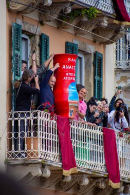 İnsanlar bir binanın balkonunda toplandı. Büyük, süslü kırmızı bir tencereyi düşürmeye hazırlanıyorlar. Kutlama sırasında olayı kameralarla çekiyorlar..
