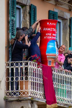 İnsanlar bir balkonda büyük, kırmızı, süslü bir nesneyi metinle tutarken görülüyorlar. Fotoğraf çekiyorlar ve bir etkinliğe katılıyorlar.