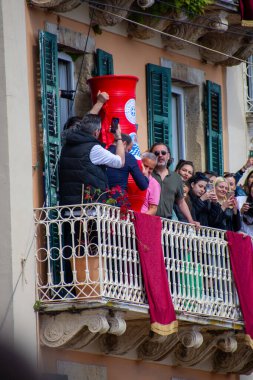 Balkondan, bir grup insan mimari detaylarla çerçevelenmiş büyük bir kırmızı konteynır fırlatırken, seyirciler olayı telefonlarıyla kaydediyor..