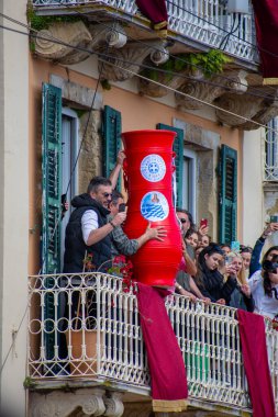 Balkon sahnesinde, balkondaki insanlar ve aşağıdaki izleyiciler telefon ve kameralarla etkinliği kaydederken büyük bir kırmızı kap yer alıyor..