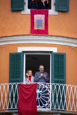 Yüksek açılı bir açıdan, bir çift yeşil panjurlu bir balkonda dikilirken görülüyor. Bina turuncuya boyanmış ve bir afişi var..