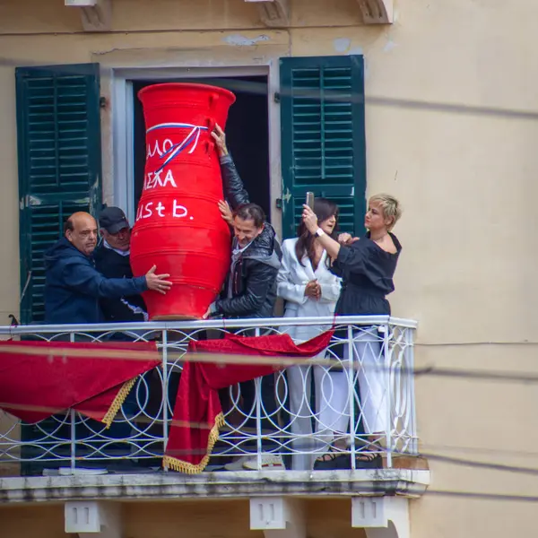 Bir grup insan birlikte çalışarak, büyük kırmızı bir vazoyu pencereden balkona taşırken, izleyiciler de o anı telefonlarla yakalıyorlar..