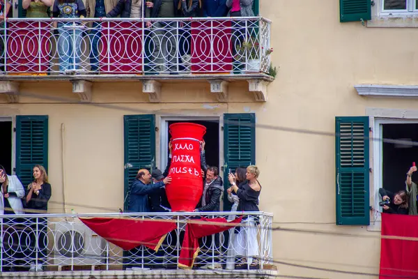 İnsanlar pencereden kırmızı bir tencere fırlatıyor. Balkonlarda insanlar var. Esrar Yunanistan 'da Paskalya için atılıyor.
