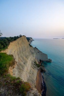 Dramatik sahil kayalıkları, sabahın erken saatlerindeki sessiz gökyüzünün altında yemyeşil bitki örtüsüyle kaplı berrak turkuaz denizine inerler..