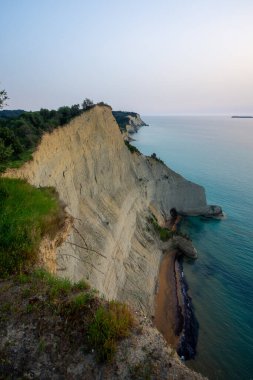 Berrak okyanus sularıyla buluşan yüksek uçurumların manzarası yumuşak bir gökyüzünün altında, yıpranmış kaya oluşumlarının tepesinde yemyeşil bitki örtüsüyle doludur..