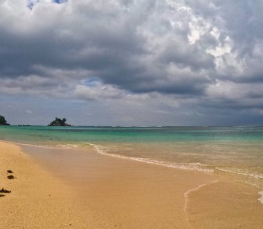 Dramatik, bulutlu bir gökyüzünün altında pitoresk bir plaj sahnesi beliriyor. Turkuaz su kumla buluşur, uzakta kayalık adalar vardır..