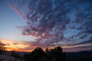 Gün batımında ağaçların ve uzak tepelerin üzerinde pembe renkli bulutlar görülür. Gökyüzü turuncudan maviliğe dönüşür.