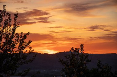 Sıcak bir günbatımı gökyüzünü turuncu ve kırmızı renklerle parlatacak, ön planda ağaçların ve tepelerin koyu gölgelerini oluşturacak..
