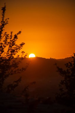Güneş ufukta batarken, altın saat günbatımı gökyüzünü turuncuya boyar. Siluetli tepeler ve ağaçlar derinlik ve kontrastlık ekler.