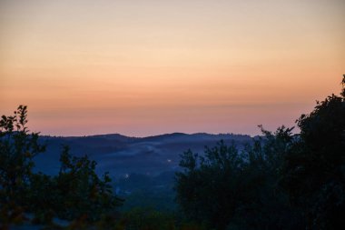 Gün ağarırken sakin bir vadi manzarası, sisle kaplı uzak tepeler. Gökyüzü ön planda yeşillik ile güzel bir renk eğimini gösterir.