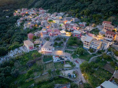 Yüksek açılı bir manzara, canlı yeşil yamaçlar ve alacakaranlıkta dolambaçlı yollarla çevrili renkli mimarisi olan huzurlu bir köyü gösteriyor..