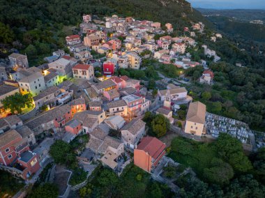 Hava manzarası, çeşitli renkli binalar ve bol ağaçlarla dolu bir yamaç köyünü görüntüler ve alacakaranlıkta manzaralı bir manzara yaratır..