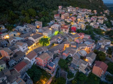 Ilık sokak lambalarıyla aydınlatılmış, ağaçların arasına yerleşmiş, hareketli evleri olan bir köyün yüksek açılı görüntüsü. Sahne huzurlu bir ortam yakalıyor..