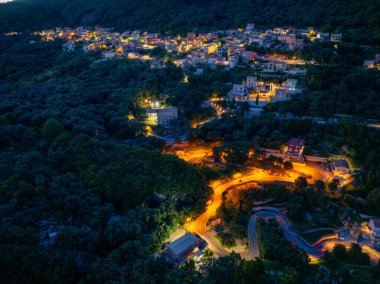Bir gece hava görüntüsü, ağaçlarla kaplı tepelerin arasına kurulmuş, yapay ışığın sıcacık parıltısıyla yıkanmış rahat bir köyü gözler önüne seriyor..