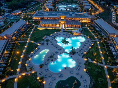 Yüksek açılı bir çekim alacakaranlıkta lüks bir otel tatil beldesini gözler önüne seriyor. Aydınlatılmış havuzlar, yemyeşil alanlar ve keşifleri davet eden yollar..