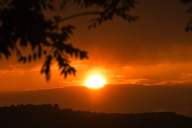Canlı bir günbatımı manzarayı sıcak renklerde yıkar, siluet ağaçları sahneyi çerçeveler. Güneşin ışıltısı bulutları delip geçiyor.