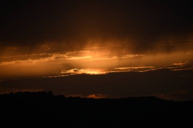 Canlı bir günbatımı bulutların arasında parlar, ufukta altın bir parıltı bırakır ve tepe boyunca ağaçların siluetini oluşturur..