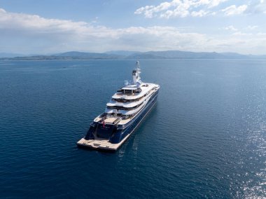 Parçalı bulutlu bir gökyüzünün altında, sakin bir denizde demirli büyük, çok katlı bir yat. Uzak dağlar ufuk çizgisinde. Huzur dolu bir sahne..