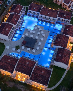 Mavi ışıklı havuzları ve terasotta çatıları olan bir otelin havadan çekilmiş görüntüsü gezginler için sakin bir alacakaranlık atmosferi yaratır..