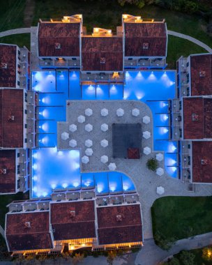 Binalar ve şemsiyelerle çevrili birden fazla havuzu aydınlatmış simetrik bir otel kompleksinin havadan çekilmiş görüntüleri hoş bir atmosfer yaratıyor..