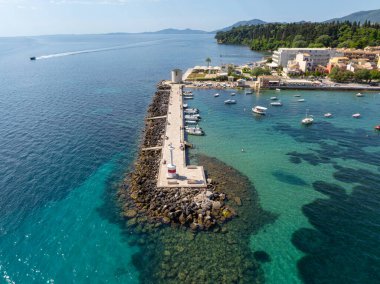 Yüksek açılı bir çekimde, kıyı boyunca tekneleri, binaları ve yemyeşil yeşillikleri olan turkuaz sulara uzanan kayalık bir iskele sergileniyor..