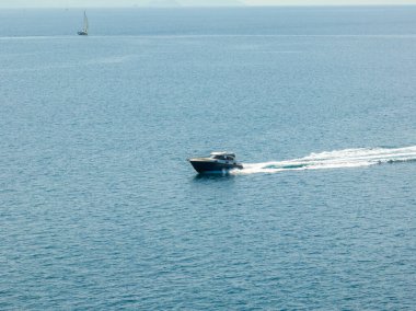 Bir motorlu tekne, uzakta görünen küçük bir yelkenliyle sakin mavi bir okyanusta hızla ilerlerken beyaz bir uyanış yaratır..