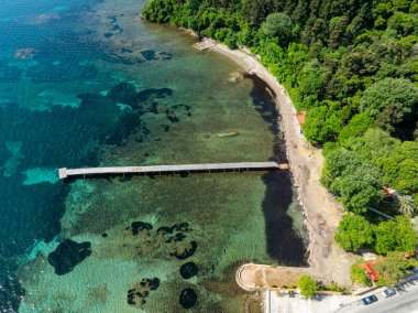 Havadan çekilen görüntülerde uzun bir iskelesi olan sakin bir sahil şeridi görülüyor. Turkuaz su, kumlu bir sahil ve yemyeşil bir ormanla tezat oluşturuyor.. 