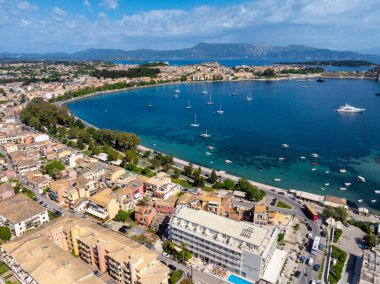 Yüksek açı, kıyı boyunca binaları olan teknelerle dolu bir körfezi gösteriyor. Arka planda dağlar yükselir, mavi bir gökyüzünün altında..