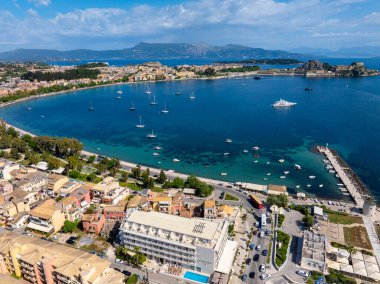 Modern bir otelin, turkuaz suların, sayısız teknelerin ve mavi gökyüzünün altındaki uzak dağların yer aldığı manzaralı bir Korfu kıyı şeridi yakalıyor..