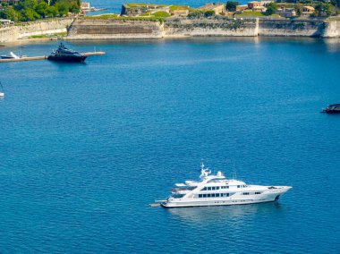 Lüks bir beyaz yat, kıyıdaki berrak mavi sularda eski bir taş yapı ve yakınındaki başka bir karanlık gemi ile geziniyor..