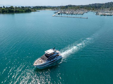Yüksek açılı bir manzara, pek çok yelkenlinin bulunduğu limanın yakınındaki turkuaz bir denizde hareket eden el değmemiş beyaz bir yatı gösteriyor..