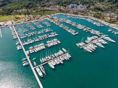 Tepeden çekimde, kıyı kasabasının arka planına ve yemyeşil yeşilliklere dayanan yat ve teknelerle dolu bir marina sergileniyor. Sakin sular..