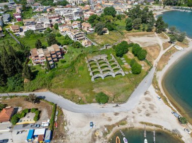 Yüksek açılı çekimde yeşil çimenler, evler, ağaçlar ve çarpıcı bir rıhtım bölgesi ile çevrili eski bir binanın kalıntıları sergileniyor..