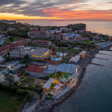 Sahilde binalar, şezlonglu kumlu bir sahil ve olay yerine sıcak ışık saçan renkli bir günbatımı görüntüsü..
