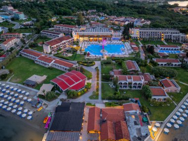 Havadan çekilmiş bir görüntüde geniş bir havuz, birden fazla bina, yemyeşil bir plaj ve şemsiyeli bir plaj görülüyor. Genel sahne çok sakin..