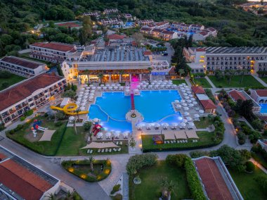 Güneş pansiyonları, binalar, yeşil peyzaj ve ağaçlarla çevrili bir yüzme havuzunun yer aldığı büyük bir tatil köyünü gösteren bir hava manzarası..
