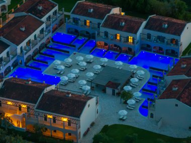 Lüks bir tatil köyünün hava manzarası, şık süitler ve şemsiyelerle çevrili mavi ışıklarla parıldayan katmanlı havuzları ortaya çıkarıyor..