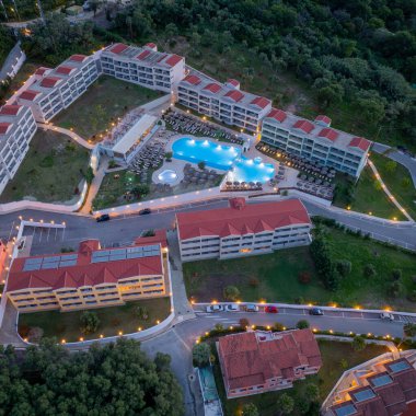 Çoklu binalar, yüzme havuzları, peyzaj bahçeleri ve alacakaranlıktaki ağaçlık alanları çevreleyen geniş bir tatil köyü kompleksinin yer aldığı bir hava görüntüsü..