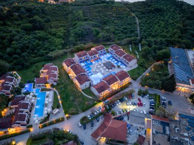 Havadan bakıldığında, havuz, mimari, verimli yeşillik ve misafirler için park tesisleri sergilenen, alacakaranlıkta lüks bir otel odası sergileniyor..