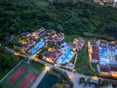 Birden fazla bina, parlak yüzme havuzları, tenis kortları ve yemyeşil ağaçların yer aldığı lüks bir tatil köyünün yukarıdan çekimleri. Otoparkta araçlar.