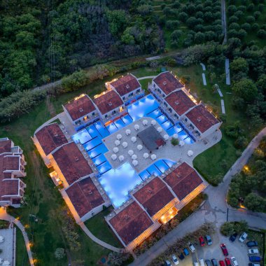 Havadan çekilen bir resimde, yeşil bir orman ve sakin bir manzaranın arasına yerleştirilmiş, mavi ışıklarla aydınlatılmış, bağlı havuzları olan bir otel tatil köyü sergileniyor..