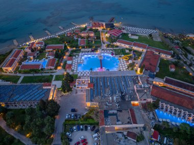 Bir hava fotoğrafı alacakaranlıkta geniş bir yüzme havuzu, geniş binalar ve resimli bir sahil şeridi içeren geniş bir tatil köyünü yakalar..