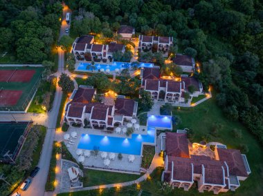 Yüksek bir manzara, mimariyi doğal çevreyle harmanlayan bir tatil beldesi gösteriyor, aydınlatma havuzları, kırmızı kiremitli çatılar ve alacakaranlıktaki patikalar.