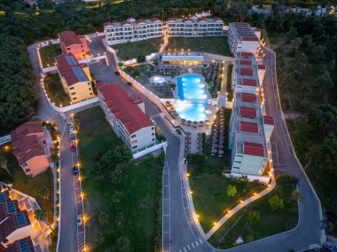 Yüksek açılı çekim, kırmızı çatıları olan binaları gösteriyor. Bir otelin havuz alanını çevreleyen yollar ve alacakaranlıkta yeşil alan manzarası var..