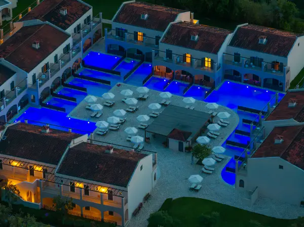 Lüks bir tatil köyünün hava manzarası, şık süitler ve şemsiyelerle çevrili mavi ışıklarla parıldayan katmanlı havuzları ortaya çıkarıyor..