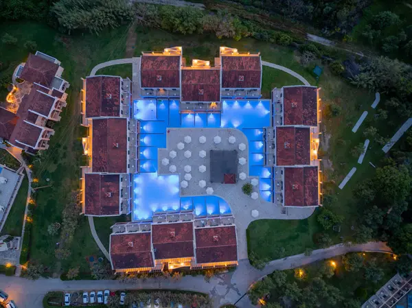Havadan çekilen bir görüntüde, alacakaranlıkta aydınlatılmış iyi tasarlanmış bir otel kompleksi görülüyor. Mavi yüzme havuzunu çevreleyen kırmızı kiremitli çatılar var..