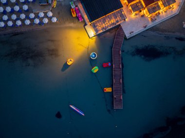 Yüksek açı karanın suyla buluştuğu kıyı şeridini gösteriyor. Tekneler gün batımında ya da şafakta, tahta bir iskelenin yanında dinlenir..