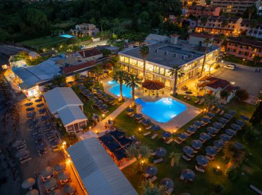 Alacakaranlıktaki deniz manzaralı bir tatil beldesi, parlayan yüzme havuzları, şemsiyeli şezlonglar ve binaların etrafındaki yemyeşil bitki örtüsü..