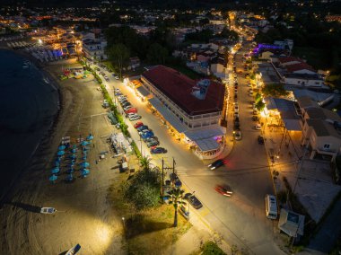 Bu hava görüntüsü, gece vakti canlı bir kıyı kasabasını yakalıyor. Sahnede şemsiyeli bir plaj, aydınlatılmış sokaklar ve mimari yer alıyor..