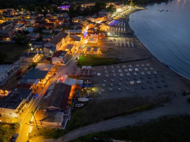 Bir gece hava görüntüsü, sahil boyunca uzanan ve binaları parlak ışıklandırılmış ve kumsalda sıralanmış plaj sandalyeleri olan bir köy gösteriyor. Deniz sakin..